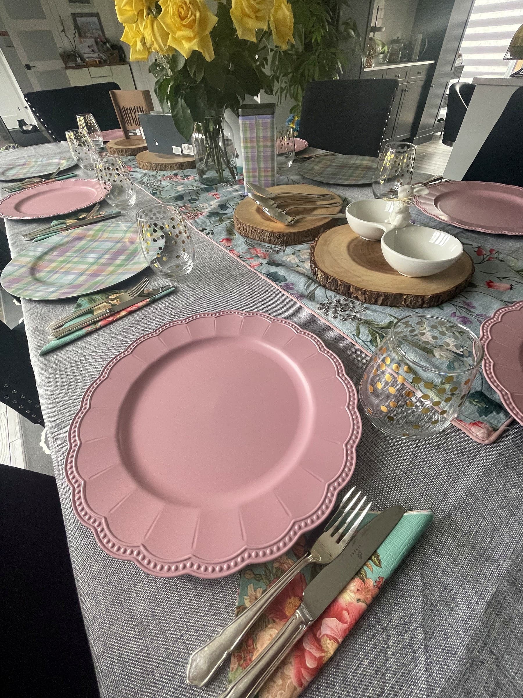 The dining table laid for Easter: pink scalloped chargers, plaid placemats, cut glass and a vase of yellow roses down the middle